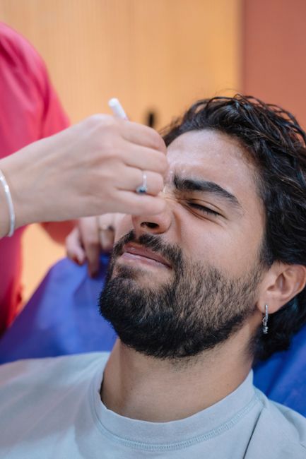 A person with dark curly hair and a beard is receiving a vaccination or injection in their upper arm from a healthcare professional wearing a red uniform. The individual is seated indoors in a room with wooden paneling in the background, and their eyes are closed as the procedure is performed. The healthcare worker's hand, visible in the image, holds a syringe and is wearing a latex glove, while the person being vaccinated appears relaxed. This scene is unrelated to house removals or moving services and does not depict any furniture, boxes, or relocation equipment. The image primarily reflects a medical or health-related context rather than home relocation or furniture transport.