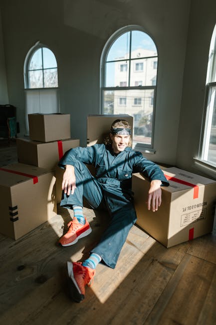 A young man sitting on the wooden floor inside a residential room during a home relocation, surrounded by packed cardboard boxes sealed with red packing tape. The boxes are stacked and arranged around him, indicating an ongoing packing process. The room has large, arched windows letting in natural daylight, with a view of neighboring buildings outside. The man, dressed in casual clothing including a denim jacket, dark pants, and bright orange running shoes, appears relaxed and engaged, possibly taking a break during the packing and moving procedure. This scene captures the preparatory stage of furniture transport or house removals, supported by the presence of moving boxes and the indoor environment. Finchley Removals’ services may include packing, loading, and home relocation within the Finchley area.