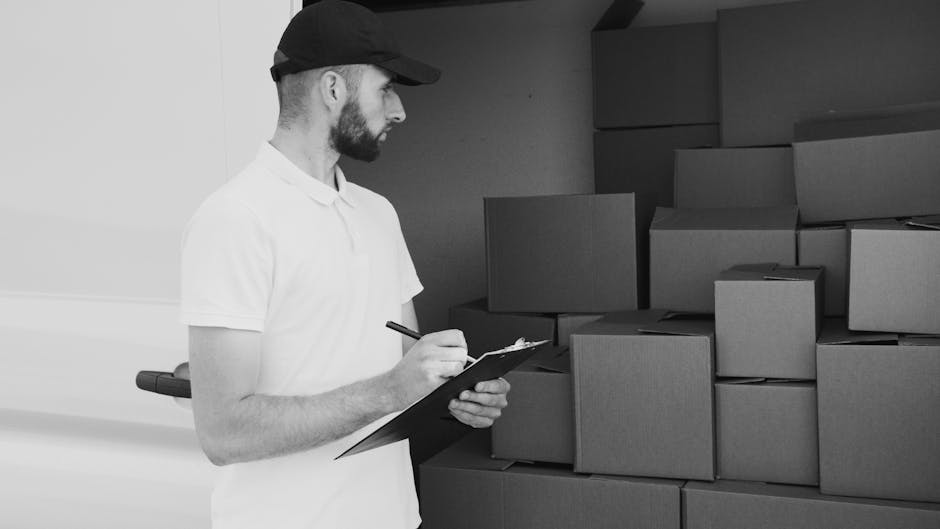 A male mover from Finchley Removals wearing a black cap and a white polo shirt is inside a warehouse or storage area, observing and taking notes on a clipboard. He is surrounded by stacks of uniformly sized cardboard boxes, some stacked on top of each other and others arranged along a wall, ready for home relocation or furniture transport. The boxes are sealed with brown packing tape and are made of corrugated cardboard, suitable for packing personal belongings during packing and moving processes. The lighting in the environment is even, highlighting the boxes and the mover’s focused expression as part of the loading process. The scene captures the careful planning and organization involved in furniture transport or tight access moves, with the boxes positioned on a smooth, clean floor which may be part of a logistics or moving company's storage facility. Finchley Removals handles such detailed packing and transportation tasks as part of their removals service, ensuring items are well-organized for efficient home relocation.