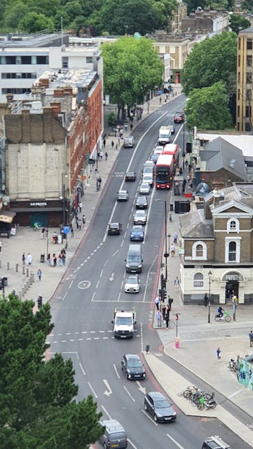 An aerial view of a busy street in Finchley with a mix of modern and traditional buildings, including a red brick structure and a white residential property with a small outdoor area. The road is lined with large, leafy green trees and has multiple lanes with cars and buses moving in both directions, some waiting at a Traffic Light junction. Sidewalks are active with pedestrians, some walking and others waiting to cross, while bicycles are parked along the pavement near a small bicycle rack. The scene captures the urban environment during daylight with clear weather, illustrating typical city street activity that Finchley Removals might encounter during home relocation or furniture transport processes involving street loading and unloading.