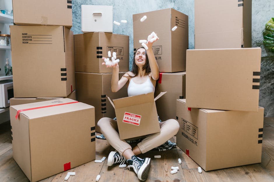 A young woman with long brown hair, dressed in casual clothing, is seated on the wooden floor inside a home surrounded by numerous unpacked cardboard boxes of various sizes. Some boxes are sealed with red packing tape and labeled with black text, while others are open, revealing packing materials such as white foam peanuts. She is smiling and actively throwing foam peanuts into the air, indicating a packing or unpacking process related to house removals. The setting appears to be a room with light blue painted walls, a small potted plant visible on the right, and a countertop with a microwave and other kitchen items on the left. The scene reflects the logistics of packing and moving, core elements of home relocation services provided by companies like Finchley Removals, emphasizing the careful handling and organization involved in furniture transport and packing during a house move.