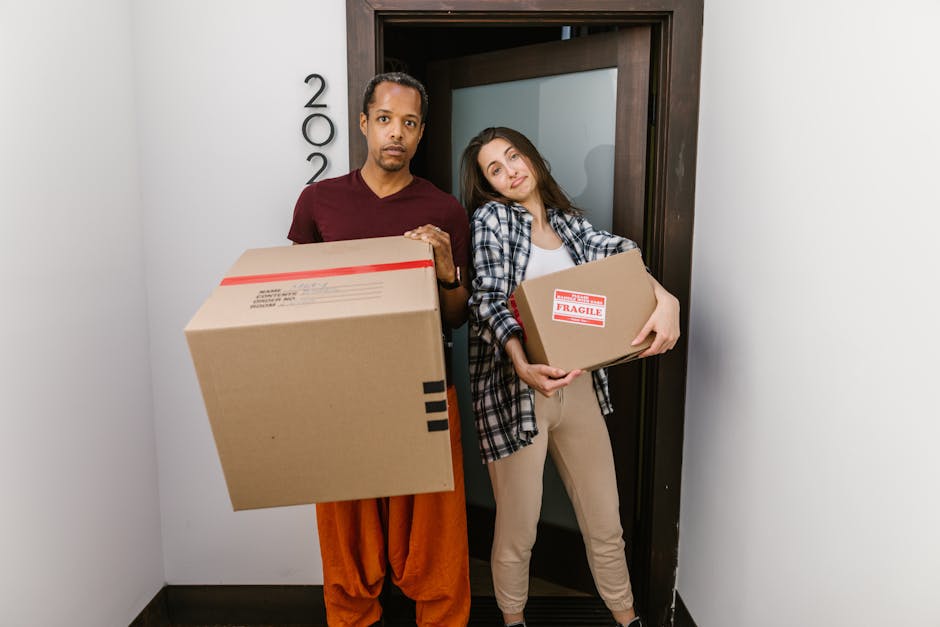 A man and a woman stand inside a property near a doorway, each holding a cardboard box secured with packing tape. The man is wearing an orange pair of pants and a maroon T-shirt, while the woman is dressed in beige trousers and a checkered shirt. The woman’s box has a red and white 'FRAGILE' label, indicating careful handling during packing and moving processes. The scene appears to be part of a home relocation or furniture transport operation, with both individuals preparing for a move and demonstrating lifting and carrying actions. The interior environment is well-lit, with a modern doorframe visible behind them. This image reflects the process of loading boxes as part of a house removals service, such as those provided by Finchley Removals, supporting effective packing, loading, and transportation in an organized manner for relocations in or around the N12 postcode area.