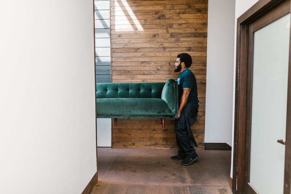 A man wearing a navy blue shirt and dark trousers is inside a residential property, carrying a green upholstered couch with a tufted design and wooden legs. The couch is being moved through a doorway, with the man's hands gripping the arms of the furniture. The room features a wooden slat wall behind him, and the floor is carpeted with a neutral tone. The doorway has a dark wood frame, and to the right, there is a partially visible glass door or window. Natural light enters the space, illuminating the scene. This image illustrates the process of furniture transport during a home relocation, with the furniture being carefully carried outside or into a different room, supported by a professional team such as Finchley Removals, who specialize in residential removals and furniture lifting services.
