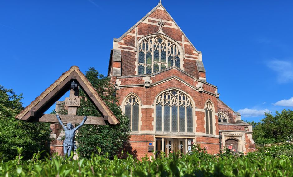 A tall, historic red-brick church with large pointed stained glass windows and decorative stone tracery, set against a bright blue sky with a few wispy clouds. In the foreground, there is a wooden churchyard cross with a crucifix, featuring a blue-tinted statue of Jesus with arms outstretched, placed on a wooden frame with an 'INRI' sign mounted above. Surrounding the church and cross, there are green trees and well-maintained shrubbery, and the image appears to be taken during daylight. This scene reflects the exterior environment of a religious building, which may be relevant in the context of house removals or moving logistics involving the transportation of large or delicate items such as church furniture or religious artifacts, with Finchley Removals providing specialised services for such relocations.